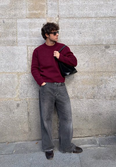 Suéter granate, vaqueros anchos grises, bolso negro. De pie contra una pared de piedra, con gafas de sol, postura relajada y manos en los bolsillos.
