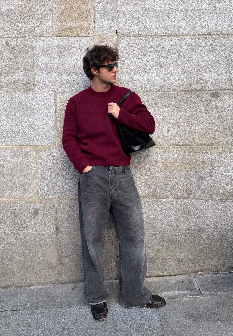 Pull marron, jean gris large, sac noir. Debout contre un mur de pierre, portant des lunettes de soleil, posture détendue et mains dans les poches.