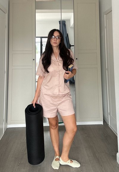 Camisa de manga corta rosa claro y pantalones cortos a juego, sosteniendo una esterilla de yoga negra y un libro; calcetines claros con tiras cruzadas. Fondo de interior moderno.