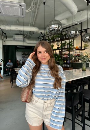 Camisa de rayas de manga larga en azul claro y blanco, combinada con pantalones cortos color crema. Bolso marrón con estampado. Entorno interior con una barra y plantas.