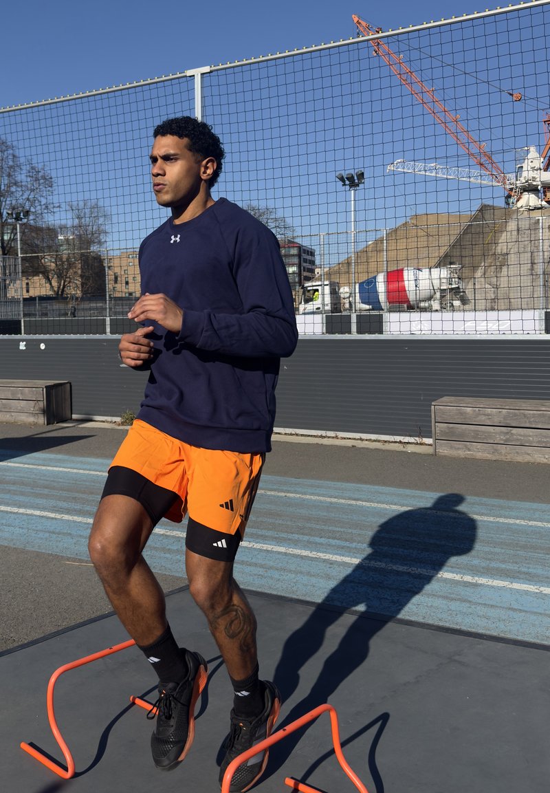 Mann i svarte joggesko, oransje shorts og marineblå genser som hopper over oransje hinder på en utendørsbane med industribakgrunn.