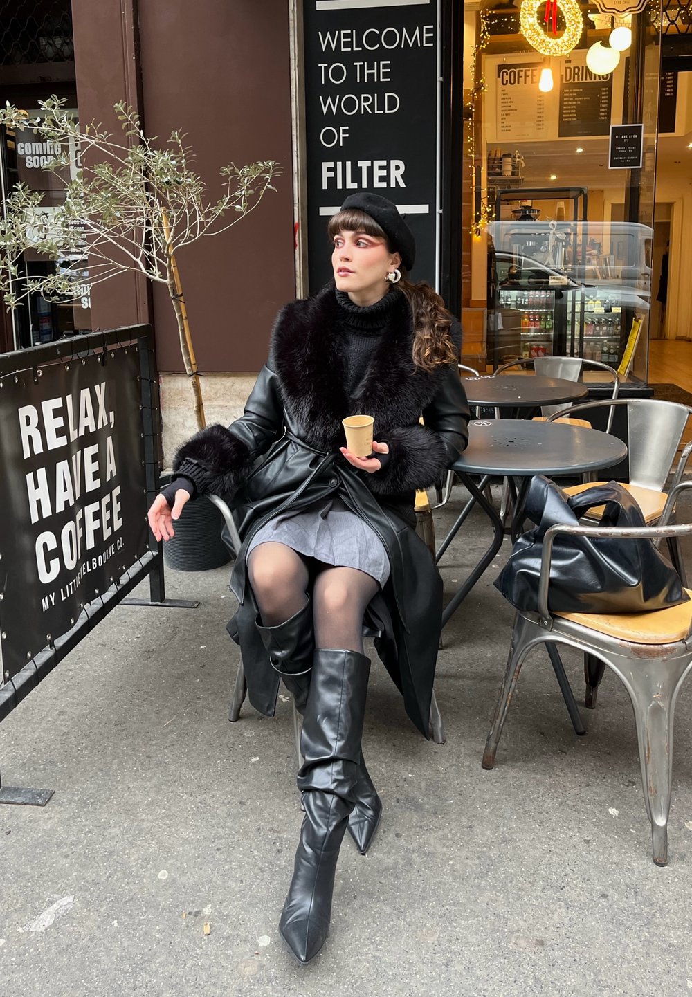 Manteau en cuir noir avec col en fourrure, jupe grise et bottes noires pointues montant jusqu'aux genoux. Tenant une tasse de café, assise à une table en métal.