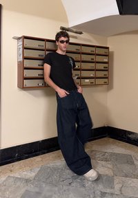 Must t-särk, lahtised tumedad denim teksad ja beežid sussid. Seisab posti sortimise seadme kõrval, millel on metallkuipe.