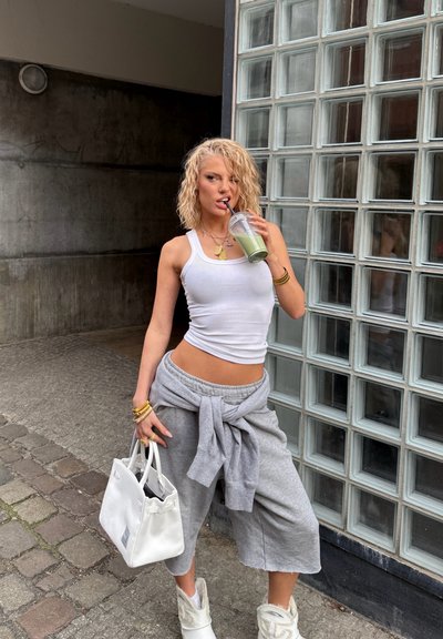 Top blanco de tirantes, pantalones cortos gris oversized, bolso blanco y botas de tobillo forradas de pelo; sosteniendo un batido verde en un vaso transparente con una pajita.