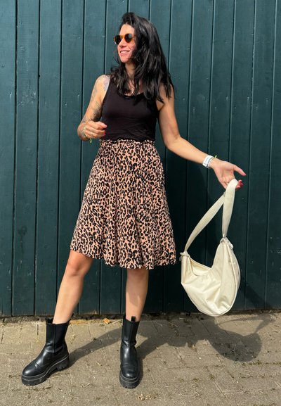 Top negro sin mangas, falda de estampado de leopardo en rosa y negro, botines gruesos negros y un bolso de hombro color crema contra un fondo de madera verde azulado.