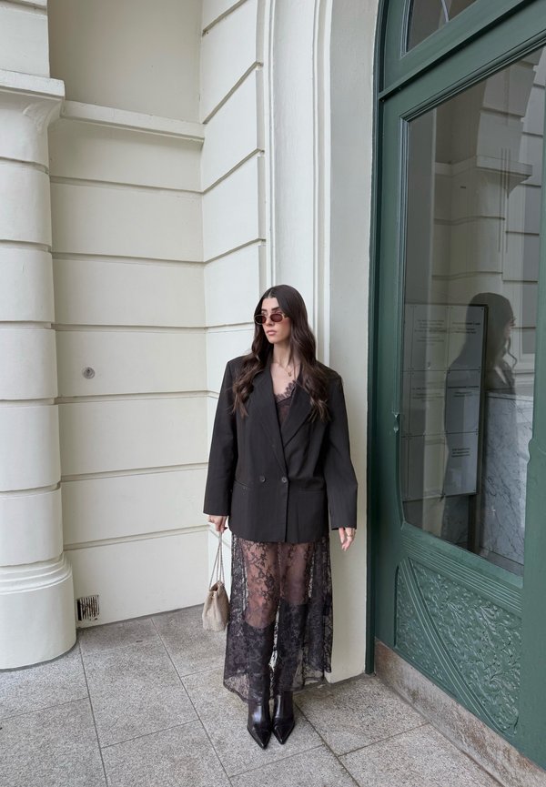 Kvinde iført sort oversized blazer, gennemsigtig sort blonderkjole, sorte støvler, solbriller, holder en beige taske, stående ved en hvid bygning med grøn dør.