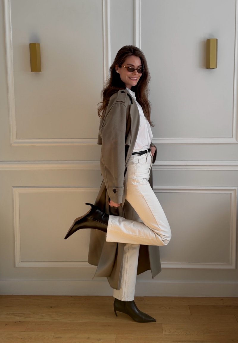 Femme portant des lunettes de soleil, un trench-coat beige, un pantalon blanc et des bottes noires à talons, levant une jambe en souriant devant un mur à panneaux.