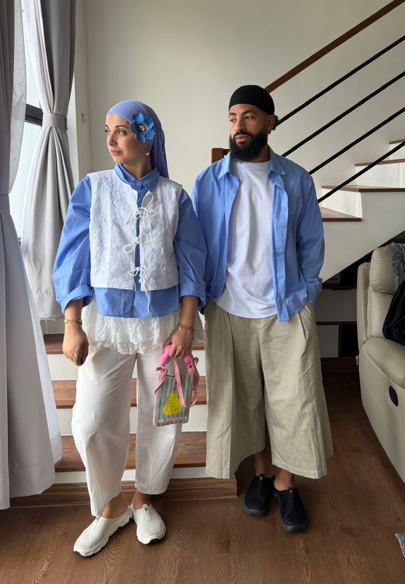 Deux personnes se tiennent près des escaliers ; la femme porte un foulard bleu avec une fleur, une chemise bleue, un gilet blanc, un pantalon blanc, tenant un sac rose ; l'homme porte une casquette noire, une chemise bleue et un pantalon large beige.