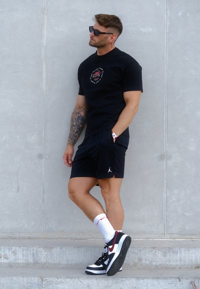 Camiseta negra de manga corta con un gráfico central, combinada con pantalones cortos deportivos negros y zapatillas negras y blancas con acentos rojos. Calcetines blancos visibles.