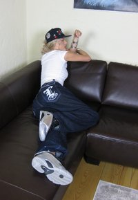 Farkut, joissa on valkoinen logo taskussa, valkoinen t-paita ja musta lippalakki. Hopeiset ja mustat lenkkarit. Istuu tummanruskealla nahkasohvalla.