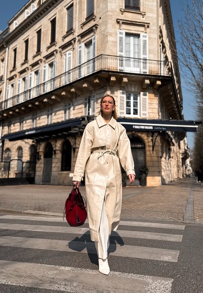 Abrigo de imitación de cuero beige claro con cintura ceñida, pantalones blancos y botas blancas de punta. Llevando un bolso tipo cubo rojo, caminando por un paso de peatones.
