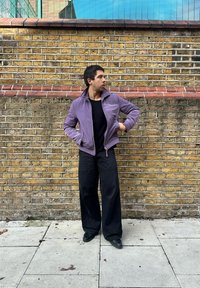 Violetti bombaripuku, jossa on korkea kaulukset, vetoketjusuljenta ja resorit hihansuissa, yhdistettynä löysiin mustiin housuihin ja mustiin kenkiin, seisomassa tiiliseinää vasten.