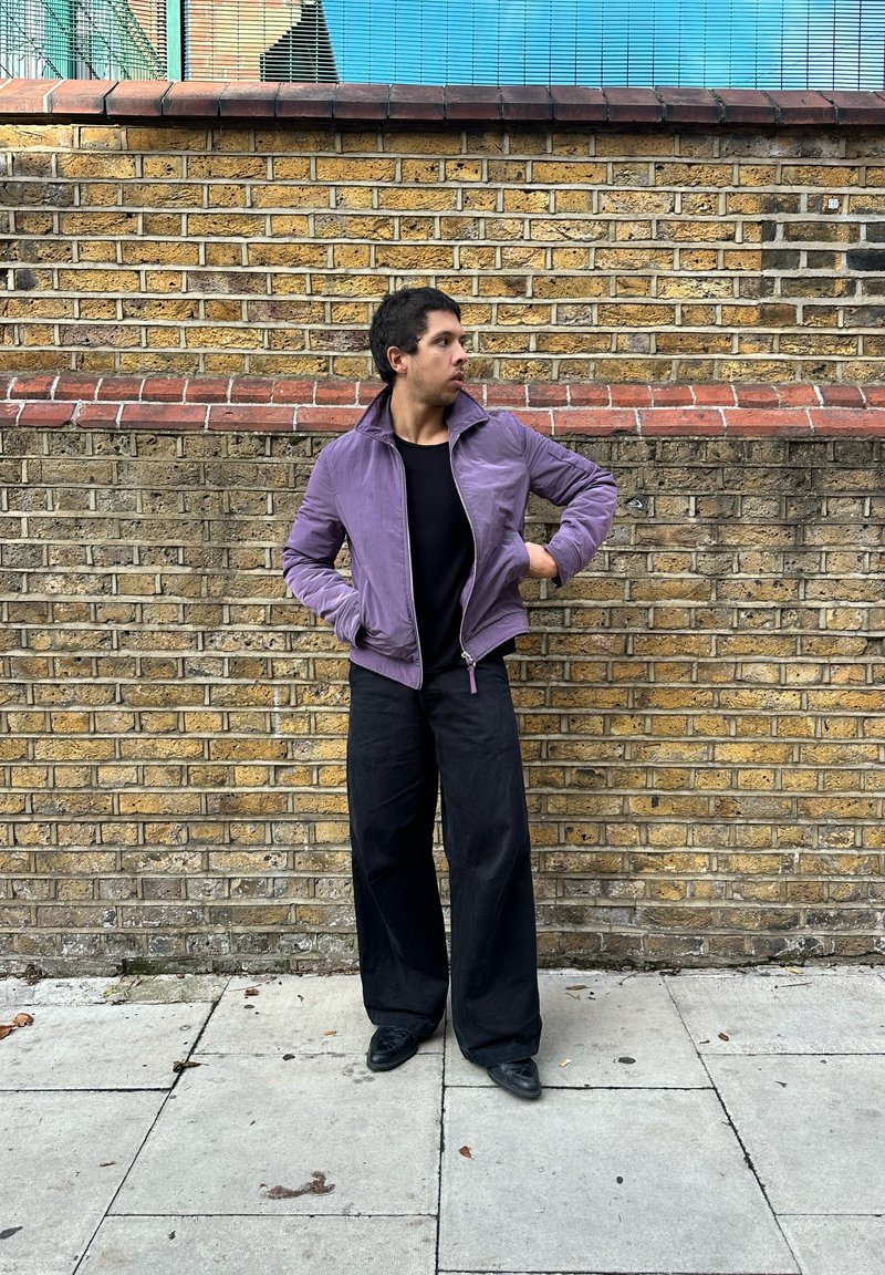 Violeta bombre jaka ar augstu apkakli, rāvējslēdzēja aizdari un ribveida aprocēm, kombinēta ar vaļīgām melnām biksēm un melniem apaviem, stāvot pret sarkanu ķieģeļu sienu.