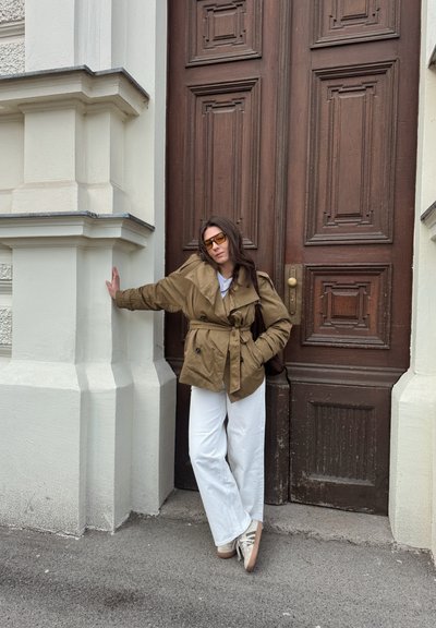 Mujer con chaqueta marrón, pantalones blancos y gafas de sol apoyada con una mano en la pared beige de un edificio junto a una gran puerta de madera oscura.