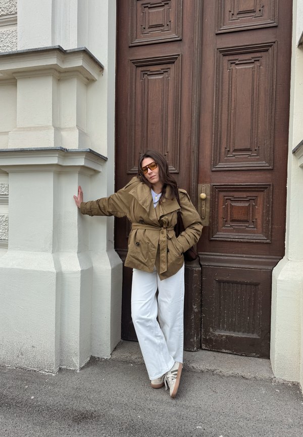 Vrouw met een bruine jas, witte broek en zonnebril die met één hand leunt tegen een beige gevel van een gebouw naast een grote donkere houten deur.