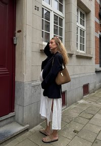 Blazer preto, vestido branco com fendas laterais, mala de ombro castanha e sandálias castanhas. Parede de pedra e porta vermelha ao fundo.