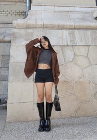 Giacca oversize in suede marrone, top corto a righe nere, pantaloni corti neri, stivali neri al ginocchio, che tiene una piccola borsa nera contro un muro texturizzato.