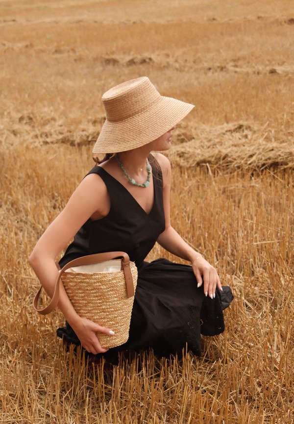 Femme portant un chapeau de paille à large bord et une robe noire, assise dans un champ sec et doré, tenant un sac panier tressé avec une lanière en cuir.