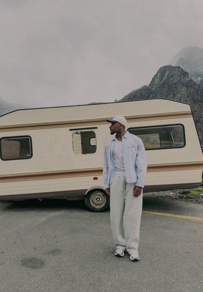 Hombre con gorra y ropa informal está junto a una autocaravana de color crema con montañas de fondo en un día nublado.