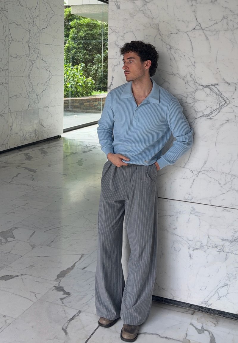 Man in light blue sweater and gray pinstripe pants leans against white marble wall inside a building with marble flooring.