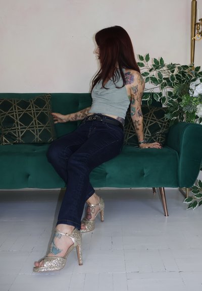 Sandalias doradas brillantes con tacón, punta abierta y correa en el tobillo. Jeans azul oscuro y una blusa sin mangas gris brillante, sentada en un sofá de terciopelo verde.