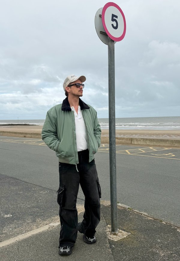 Hombre con chaqueta verde, pantalones cargo negros y gorra, de pie junto a un poste con una señal de límite de velocidad que muestra el número 5 junto al mar.