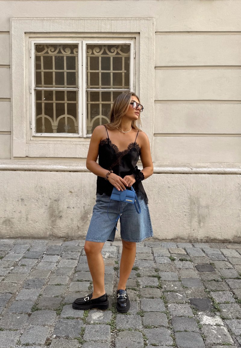 Camisole nera con pizzo, pantaloncini di jeans e mocassini neri. Tiene in mano una piccola borsa azzurro chiaro. Pavimentazione in ciottoli sullo sfondo.