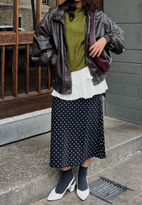 Brun skinn bomberjakke, grønn strikket genser, hvit lagdelt skjørt med volanger og svarte polkadotter, grå sokker, hvite blokkhælede sko.