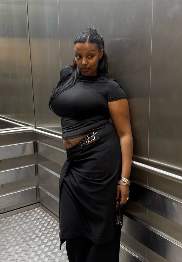 Femme aux longs cheveux se tient dans un ascenseur en métal, portant un top court noir, une jupe noire avec ceinture, tenant des lunettes de soleil dans une main.