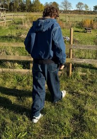 Kapucnis farmerdzseki, oversized fazon. Szellős kék farmernadrággal párosítva. Fehér sportcipők. Zöld fű és fából készült kerítés háttér.