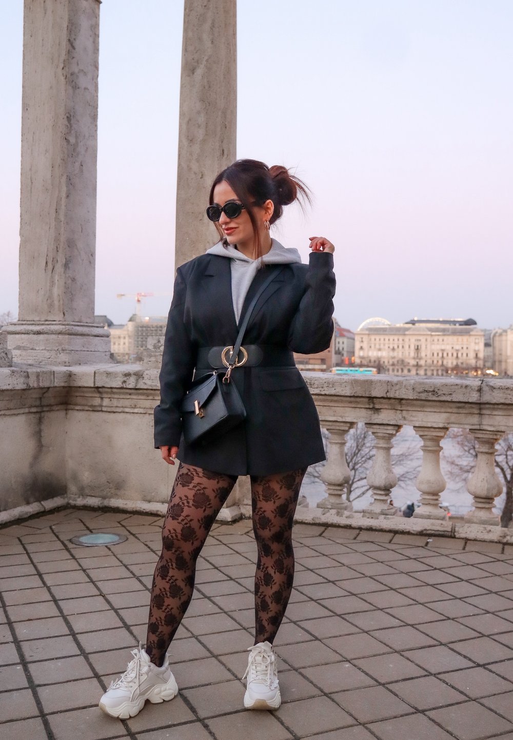 Blazer preto ajustado com cinto, hoodie cinza, collants com padrão floral e ténis brancos. Óculos de sol escuros e uma pequena bolsa destacam o visual.