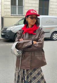 Hnědá kožená oversized bunda, červený šátek a baseballová čepice. Stříbrná kabelka s řetízkovým detailem, pod ní kostkovaná sukně. Sluneční brýle dotvářejí vzhled.