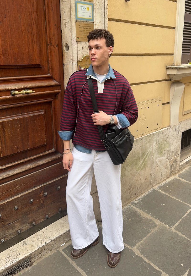 Νέος άνδρας με κόκκινη ριγέ μπλούζα πάνω από τζιν, λευκό παντελόνι φαρδιάς γραμμής, καφέ παπούτσια και μαύρη τσάντα ώμου στέκεται σε αστική οδό κοντά σε ξύλινη πόρτα.
