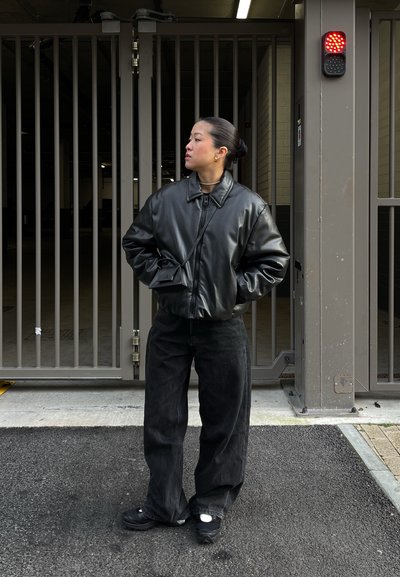 Chaqueta bomber de cuero negro, pantalones de pana negra de corte relajado y zapatillas oscuras. De pie fuera de una zona cerrada con una luz roja.