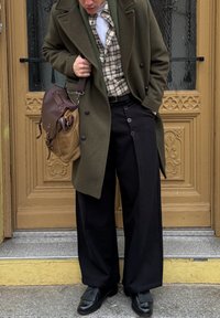 Oliwkowo-zielony dwurzędowy płaszcz na koszuli w kratę, czarne szerokie spodnie i czarne buty. Brązowa torba na ramię z metalowymi dodatkami.