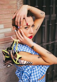 Sandales à talons hauts avec imprimé zèbre et lanières vert néon. Modèle portant un haut à carreaux bleu pose avec des couleurs d'ongles audacieuses et des tatouages visibles.