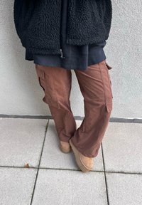 Καφέ cargo παντελόνια με πλαϊνές τσέπες, υφασμάτινη υφή, φαρδιά υφή ποδιών, σε συνδυασμό με καφέ πλατφόρμες σε πλακόστρωτη επιφάνεια. Ορατό μαύρο φλις μπουφάν.