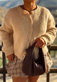 Cárdigan de punto en color beige con cuello redondo y cinco botones, combinado con una falda de cuadros y un bolso de mimbre marrón. La textura es suave y gruesa.