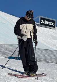 Skiingový outfit zahrnuje černobílou bundu, černé kalhoty, vzorované rukavice a černé lyžařské boty. Lyže jsou barevné s fialovým designem.