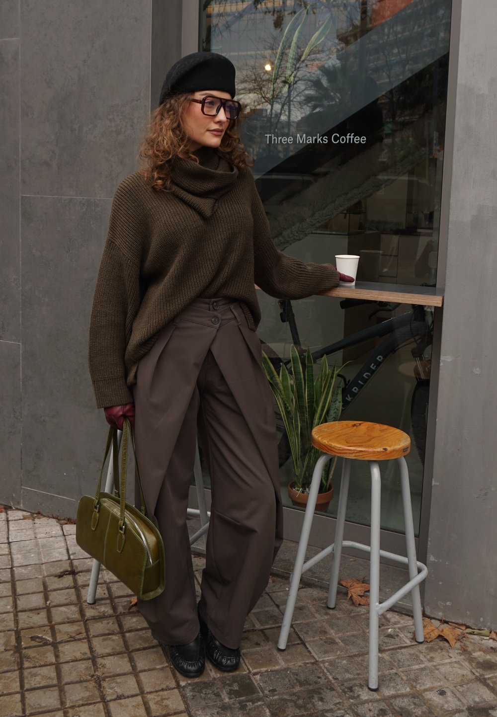 Pull tricot épais vert olive, pantalons large marron, béret noir, lunettes surdimensionnées, sac à main vert, debout devant un café.