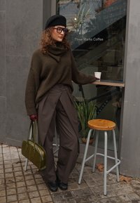 Pull tricot épais vert olive, pantalons large marron, béret noir, lunettes surdimensionnées, sac à main vert, debout devant un café.