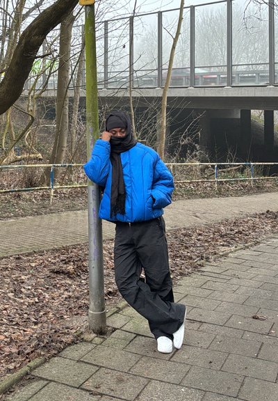 Chaqueta de plumón azul, bufanda negra y pantalones negros oversized. De pie junto a un poste, con zapatillas blancas, fondo de árboles y un puente.