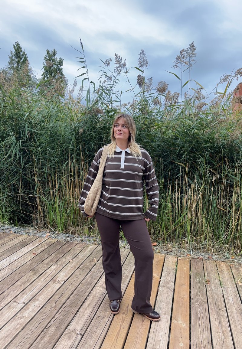 Camicia a maniche lunghe a righe marroni e bianche con colletto bianco, abbinata a pantaloni marroni e scarpe scure. Sullo sfondo si vedono piante verdi e un decking in legno.