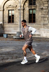 Tetoválásokkal rendelkező férfi fut egy macskaköves utcán, ujjatlan dzsekit, rövidnadrágot, fehér sportcipőt és okosórát viselve.