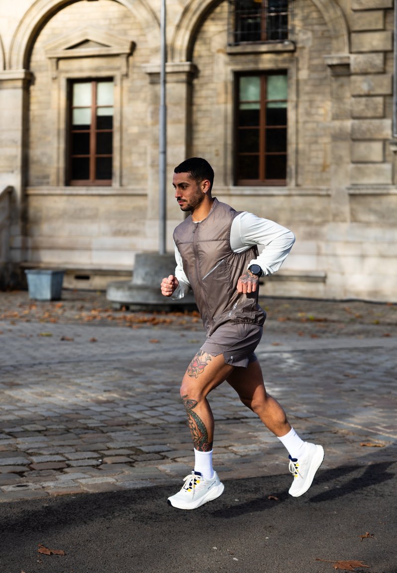 Mann mit Tätowierungen, der auf einer Kopfsteinpflasterstraße läuft, trägt eine ärmellose Jacke, Shorts, weiße Turnschuhe und eine Smartwatch.