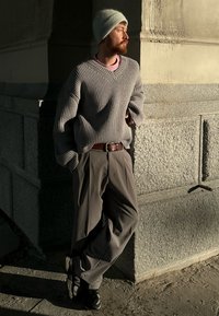 Pull gris côtelé avec manches surdimensionnées, chemise rose clair en dessous, bonnet beige, pantalon gris ample et baskets noires devant un mur en pierre.