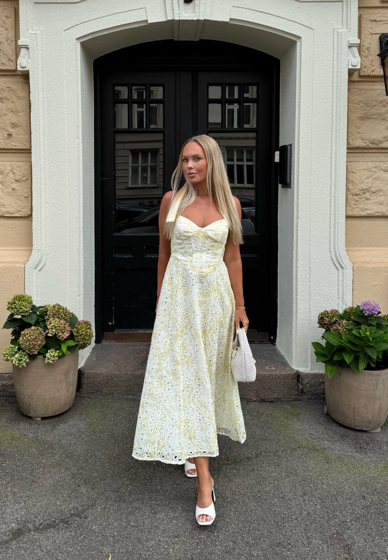Femme en robe jaune clair à œillets et talons blancs, tenant un sac à main blanc devant une porte noire encadrée par des murs beiges.