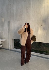Cappotto in pelliccia sintetica beige, top marrone e pantaloni sportivi marrone scuro con strisce bianche. Tiene in mano una tazza di caffè e una borsa marrone testurizzata.