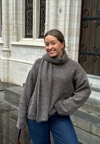 Pull en tricot texturé marron avec un col montant et une écharpe attachée, associé à un jean bleu. Un accent de chaîne en or est visible autour du cou.