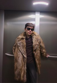 Jeune personne portant des lunettes de soleil foncées et un grand manteau en fourrure marron, s'appuyant en arrière avec une main sur une rampe métallique d'ascenseur.